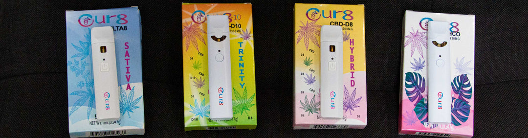 Wholesale Cur8 Delta 8, Delta 10 and THC 0 disposable vaporizers resting on their packaging on top of a textured dark background.