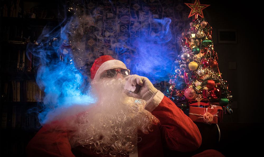 man in Santa costume sitting on chair using vaporizer and blowing smoke with Christmas tree in the background