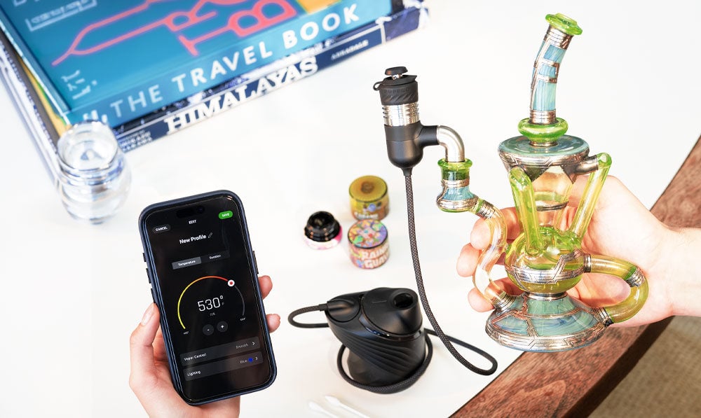 Hands holding Puffco Peak Link over white desk with waterpipe attached and Puffco app open on phone