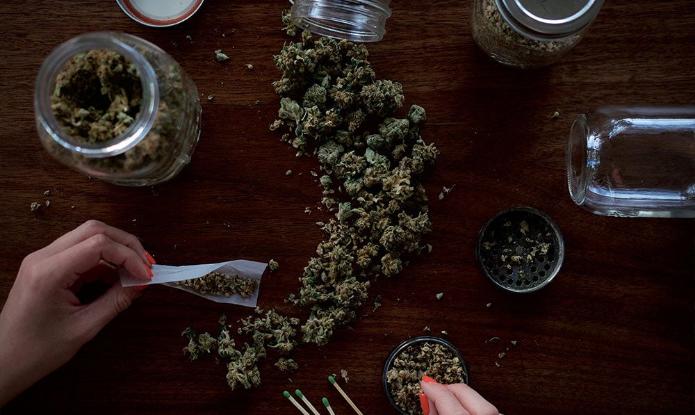 Hands rolling herb at a table with a grinder and glass jar